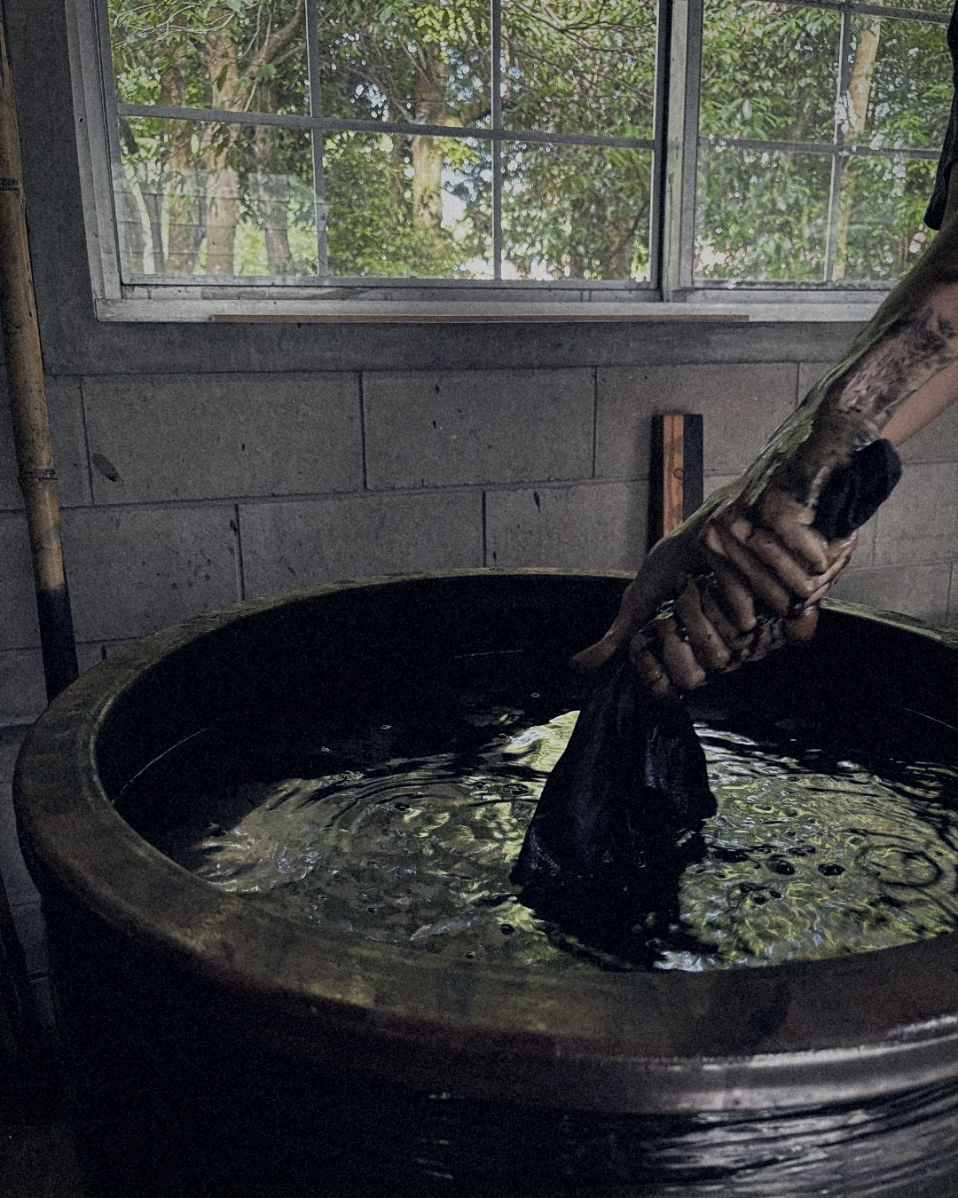 Artisan dipping cloth into a ceramic indigo vat beneath a workshop window